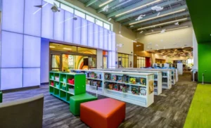 Interior library shot with shelves of books, seating and what appears to be white paper art hanging from the ceiling. It's very colorful with green and red seating.